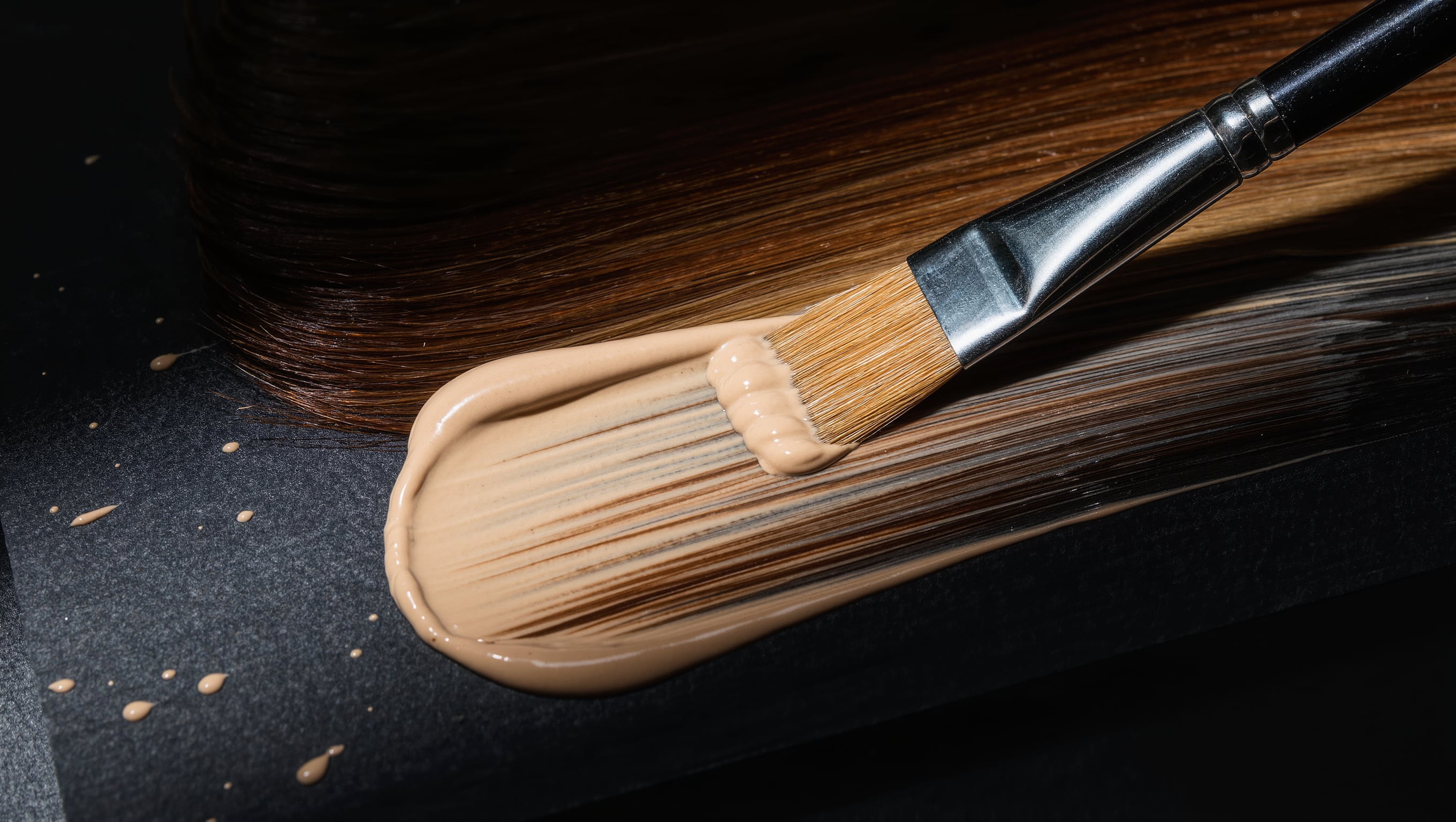 Pinsel trägt helle Blondiermasse freihand auf eine ausgebreitete Haarsträhne auf, weiches Streiflicht auf der Textur